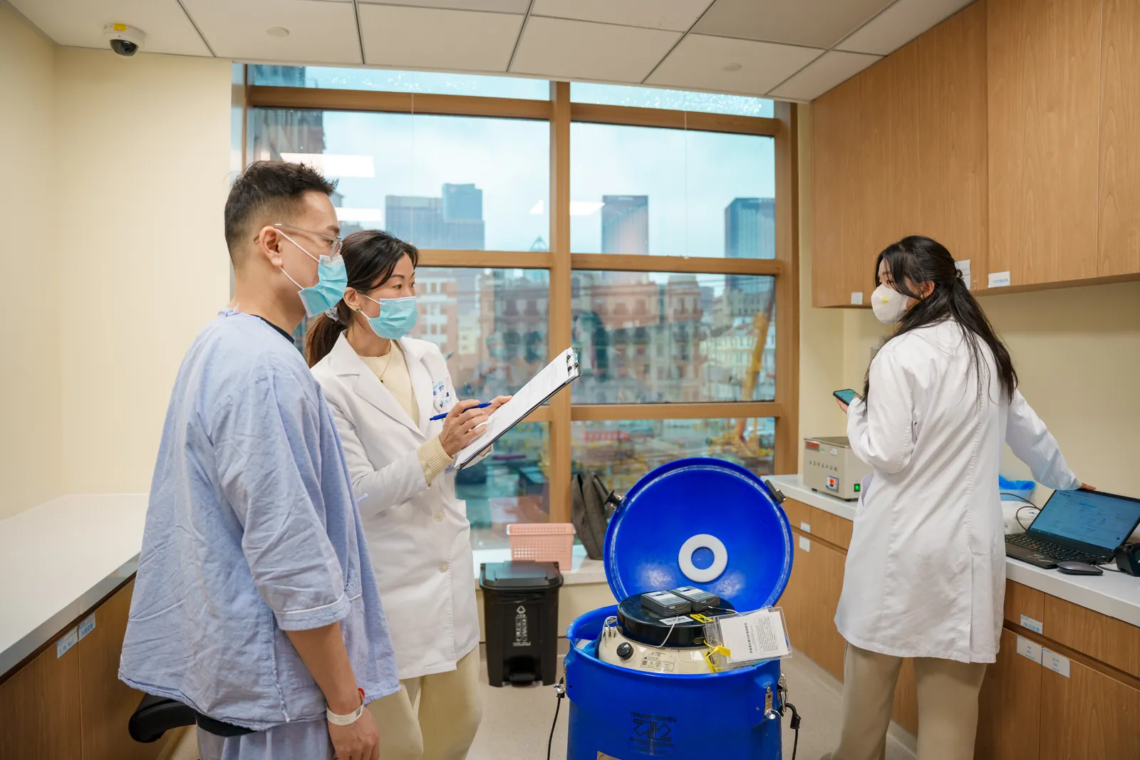 A patient and two clinicians at SinoUnited Health reviewing records beside CAR T-cell therapy equipment in a treatment room overlooking Shanghai.
