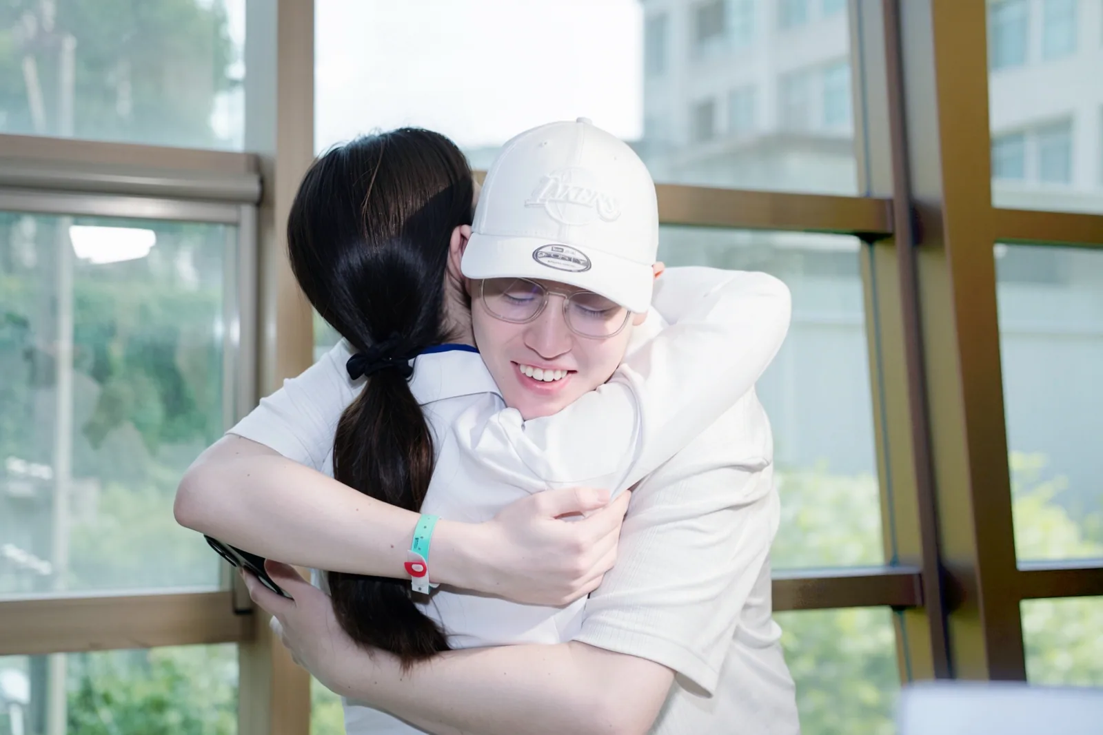 A patient in a baseball cap embracing a nurse beside the window wall of a hospital day room