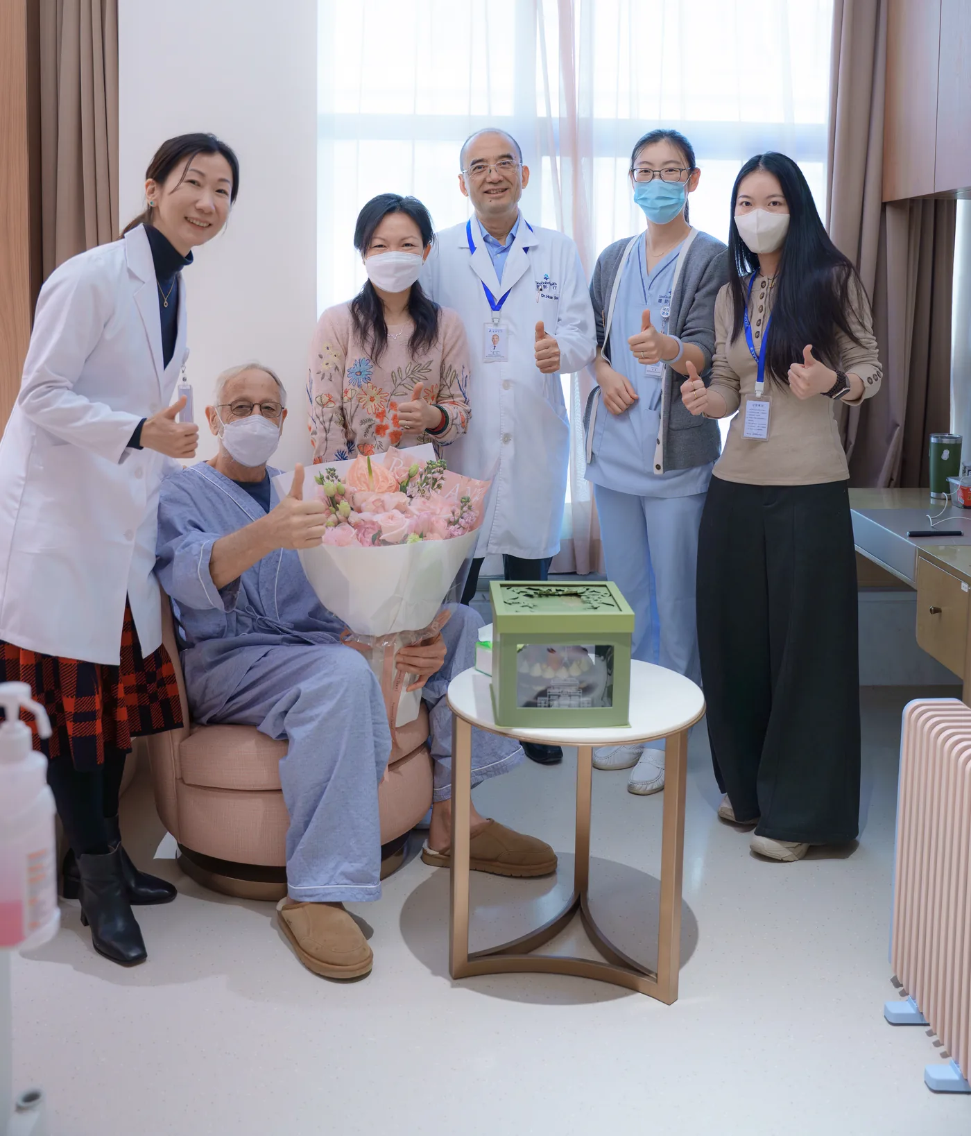 An international patient in the SinoUnited Health inpatient ward, seated with a bouquet, alongside a family member and five members of the care team
