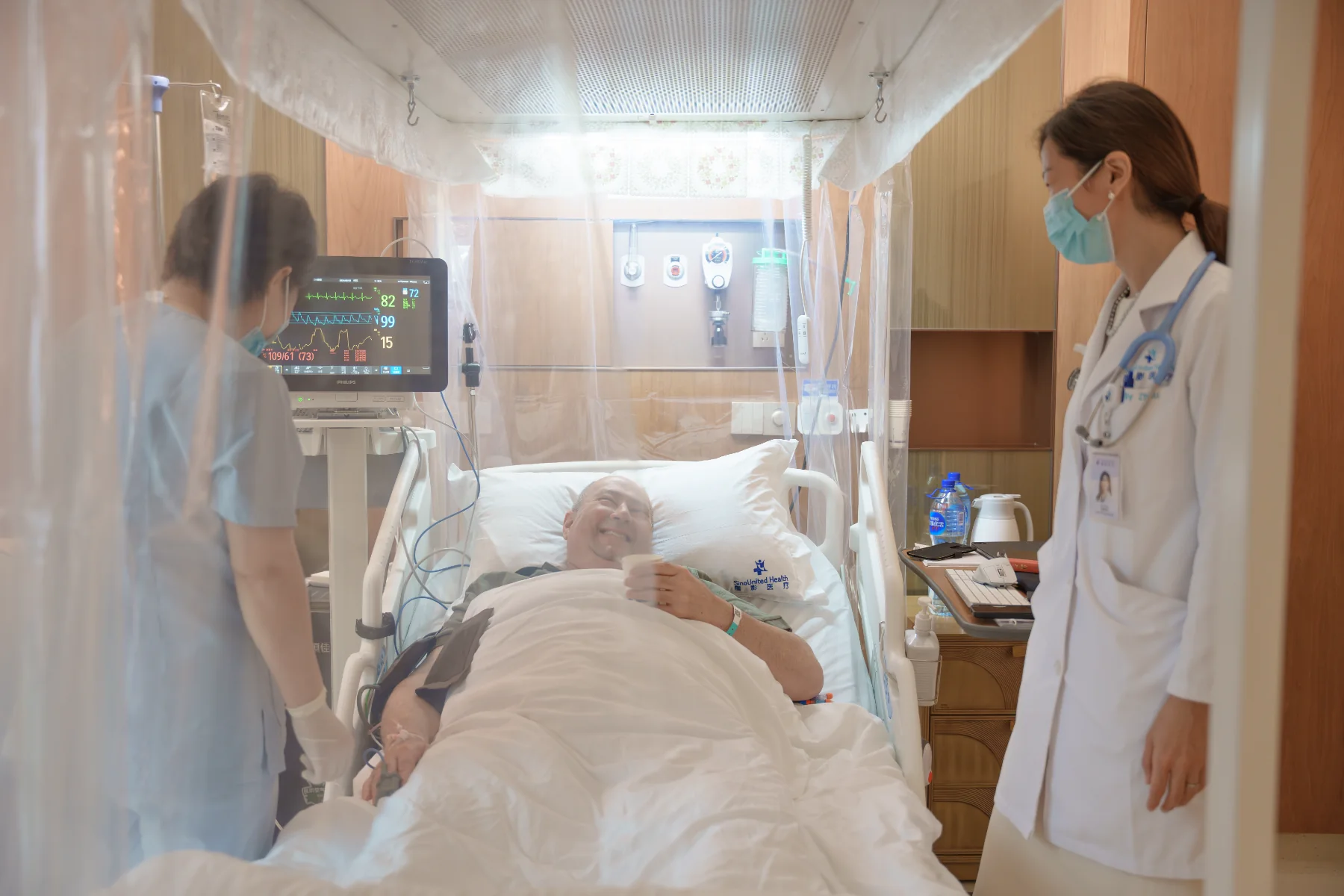 A patient rests in a private inpatient room under a laminar-flow protective canopy over the bed, with a nurse and doctor at his side, at Shanghai SinoUnited Hospital